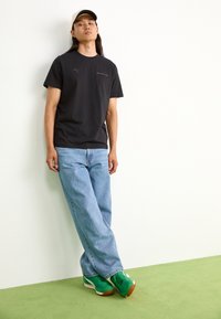 Czarna koszulka z logo Puma, jasnoniebieskie dżinsy i zielone sneakersy z brązowymi podeszwami. Stoi przy neutralnej ścianie na zielonym dywanie.