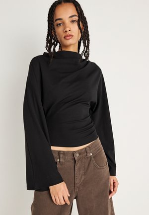 Črni dolg rokav s visokim ovratnikom in sproščenim krojem. Mehek material, gladka tekstura, v kombinaciji z rjavimi žametnimi hlačami, ki imajo podrobnosti žepov.