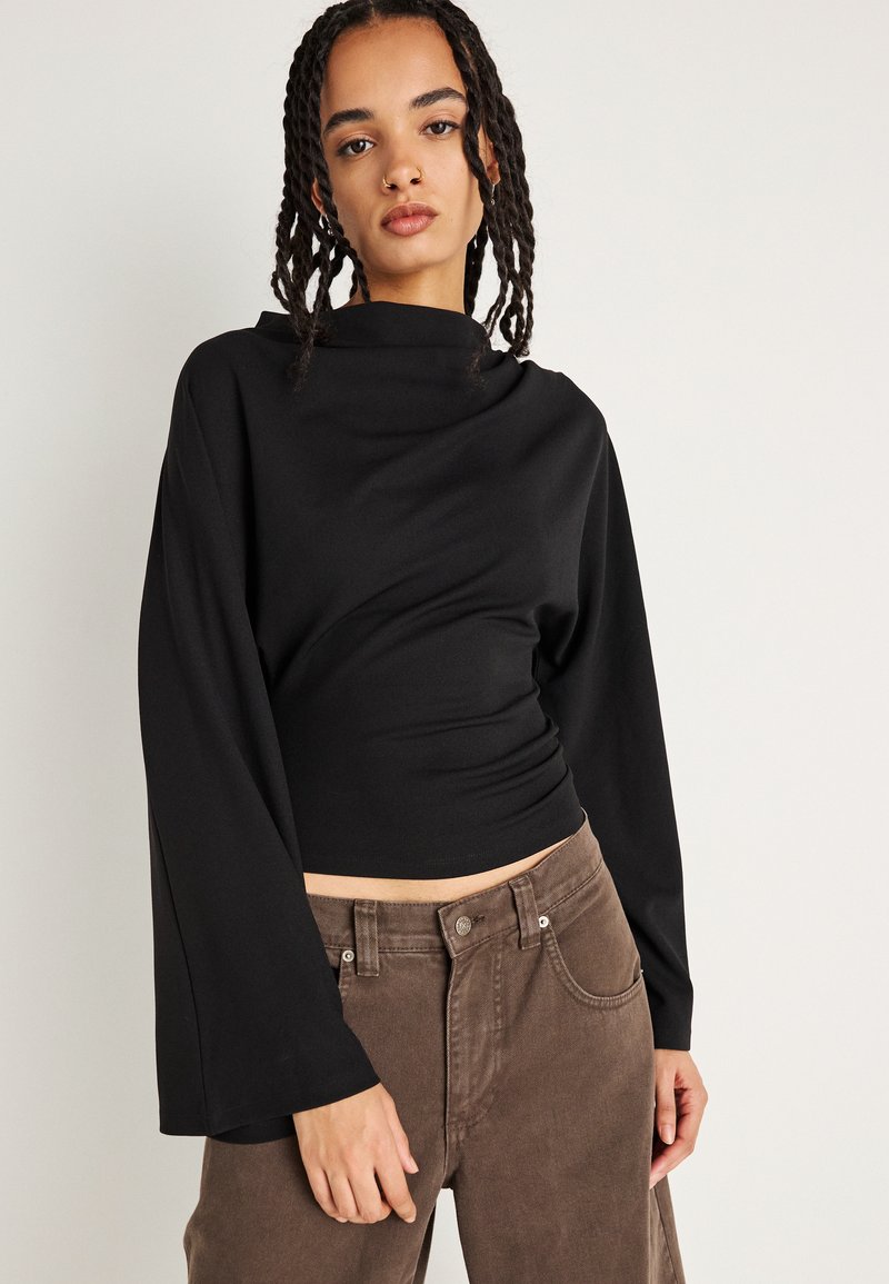 Camiseta negra de manga larga con cuello alto y corte relajado. Tejido suave, textura lisa, combinada con pantalones de pana marrones con detalles en los bolsillos.