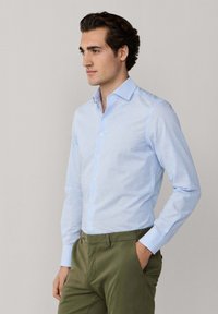 Jeune homme portant une chemise à rayures bleu clair boutonnée et un pantalon vert olive, debout avec une main dans la poche, regardant sur le côté.