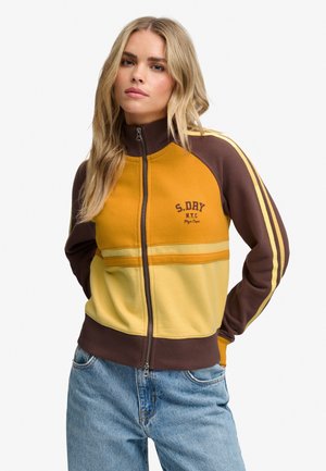 Vrouw met lang blond haar, die een bruin-gele jas met rits en blauwe spijkerbroek draagt tegen een effen witte achtergrond.