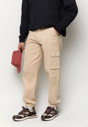 Person iført beige cargobukser, sort langærmet skjorte, brune sneakers, hvide sokker, der holder en rød kasket i venstre hånd.