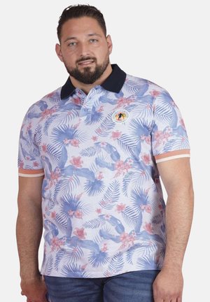 Homme portant un polo à manches courtes à motif floral avec un col noir et un jean, debout devant un fond uni.