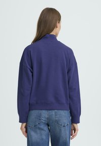 Vijoličen pulover z visokim ovratnikom, spuščeni rameni in rebrastimi manšetami, v kombinaciji z svetlo modrimi kavbojkami. Mehka bombažna tkanina.