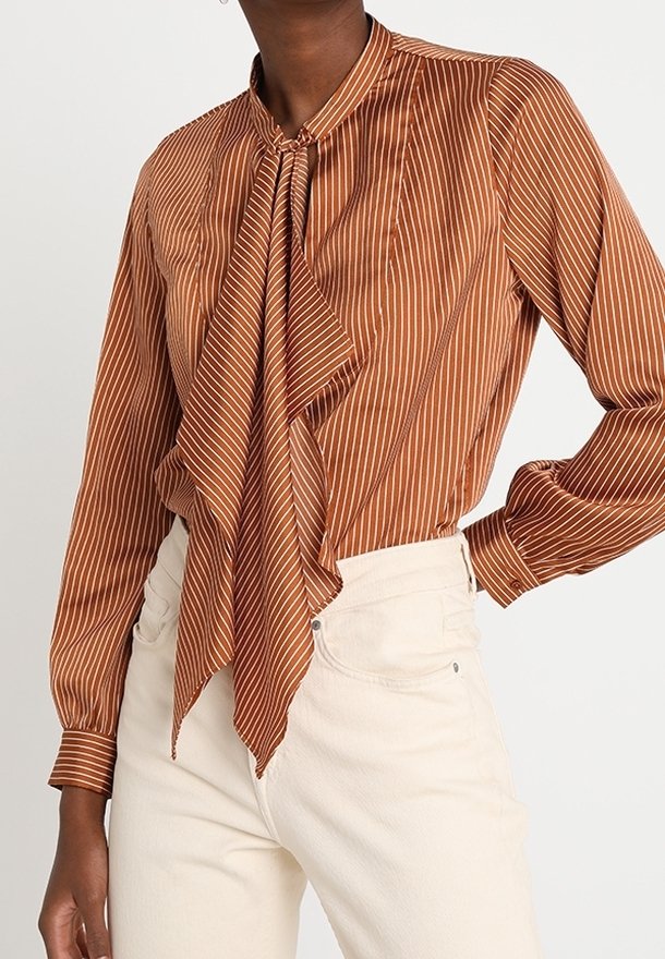 Blouse marron à manches longues avec des rayures verticales blanches et un grand nœud devant, associée à un pantalon taille haute couleur ivoire.