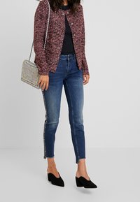 Gebreide bordeauxrode cardigan met textuur en metalen knopen, gecombineerd met aansluitende blauwe spijkerbroek met luipaardprint aan de zijkant en zwarte hakken.