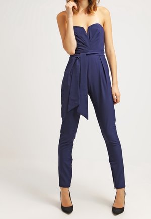 Femme portant une combinaison bleu marine sans bretelles avec une ceinture nouée à la taille et des escarpins noirs, debout devant un fond uni.