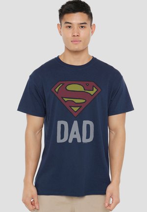 Young man wearing a navy blue T-shirt with a red and yellow Superman logo above the word "DAD" in gray letters.