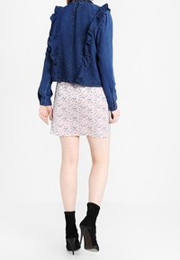 Femme portant une veste en denim bleue à volants, une mini-jupe florale rose et des bottines noires à talons hauts, se tenant de dos sur fond blanc.