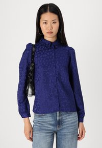 Navy blue, textured button-up shirt featuring heart patterns, long sleeves, and a classic collar. Paired with a small black shoulder bag.