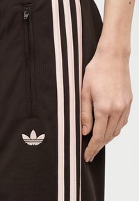 Zbliżenie dłoni obok czarnych spodni z trzema bladymi różowymi paskami, kieszenią na zamek i logo Adidas Trefoil.