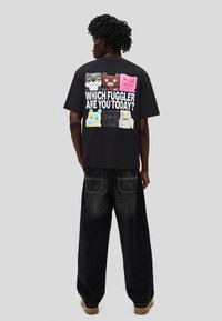 Tricou negru cu imprimeuri colorate cu personaje de desen animat pe spate, cu textul "CARE FUGGLER EȘTI ASTĂZI?" în litere albe îndrăznețe.