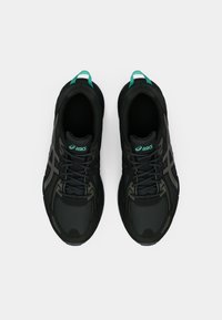 Sapatos de desporto pretos com um upper texturizado, detalhes verdes no calcanhar e no logotipo, e detalhes de marca cinzentos nas laterais.