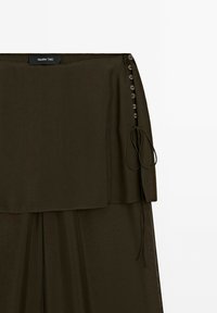 Blouse vert olive au design fluide, avec un ourlet en couches et des accents de boutons sur les côtés. Liens aux manches pour un ajustement personnalisé.