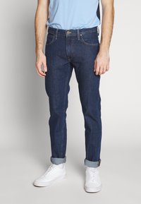 Tmavě modré džíny s úzkým střihem, sešitými kapsami, knoflíkovým zapínáním a ohrnutými lemy. Kombinované s bílými teniskami.