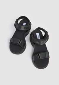 Sandalias negras con una correa texturizada que lleva la marca "PEPE JEANS". Fabricadas con materiales sintéticos, cuentan con una plantilla contorneada y suela plana.