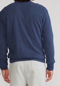 Felpa blu scuro con collo tondo in tessuto testurizzato, con maniche lunghe e un bordo in vita aderente. Indossata sopra pantaloni grigio chiaro.