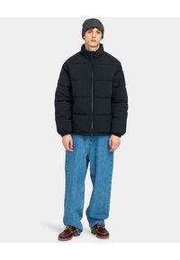 Giacca a vento nera con colletto alto e chiusura con zip, abbinata a jeans blu largo e scarpe marroni. Berretto grigio sulla testa.