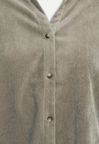 Oliwkowa koszula z korduroju z dekoltem w serek, z pięcioma ciemnymi brązowymi guzikami i miękkim, strukturalnym materiałem. Prosty design z pionowymi liniami.