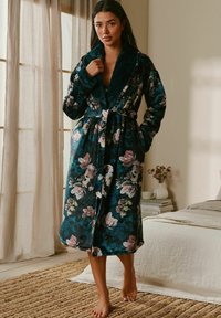 Woman standing barefoot in a bedroom wearing a dark floral robe with a plush collar, holding the collar near her neck.