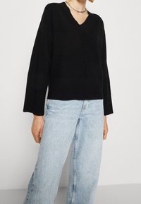 Svart, långärmad V-ringad tröja med avslappnad passform och ribbad detalj vid fållen, kombinerad med ljusblå, vida jeans.