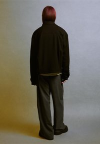 Fekete, túlságosan nagy kabát sima textúrával, laza, szürke, csíkos nadrággal párosítva. A háttér szürkéből sárgába omló gradient.