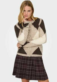 Beige kardigan s hnědými a bílými geometrickými vzory, kulatými zlatými knoflíky a měkkou texturou, doplněný tmavě kostkovanou plisovanou sukní.