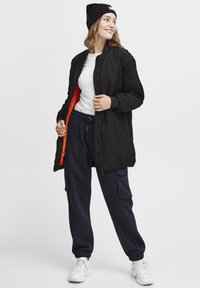 Jachetă bomber neagră cu căptușeală portocalie, tricou alb, pantaloni de trening bleumarin cu buzunare și pantofi sport albi. Modelul poartă o căciulă neagră.