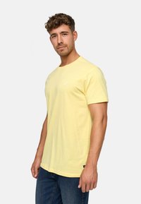 T-shirt en coton jaune clair avec des manches courtes, un col rond et un petit logo sur la poitrine. Associé à un jean en denim bleu foncé.