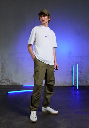 White cotton t-shirt with a small blue logo, olive green cargo pants, and white sneakers with a tan sole. Set against a textured wall.