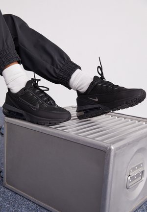 Person wearing black athletic shoes with visible air cushioning, white socks, and black pants resting feet on a metallic storage case.