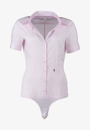 Vrouw loopt, draagt een lichtroze blouse met korte mouwen en knopen, nauwsluitende zwarte broek, zwarte open-teen hakken, houdt een beige clutch in linkerhand.