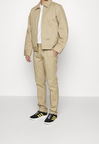 Veste et pantalon beige en tissu solide et robuste. La veste possède un col et un petit logo. Baskets noires avec des rayures jaunes.