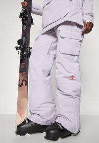 Ski oblečení s lehkými fialovými zateplenými kalhotami s cargo kapsami, doplněné černými lyžařskými botami a lyží s vázáním drženou v jedné ruce.