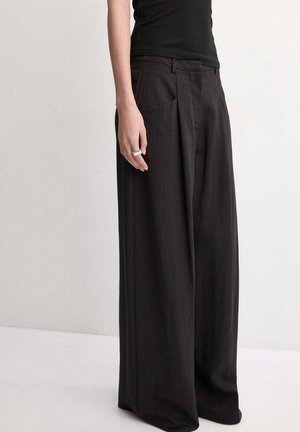 Persona con pantalones negros de pierna ancha y una camiseta sin mangas negra, con un anillo plateado en la mano izquierda.
