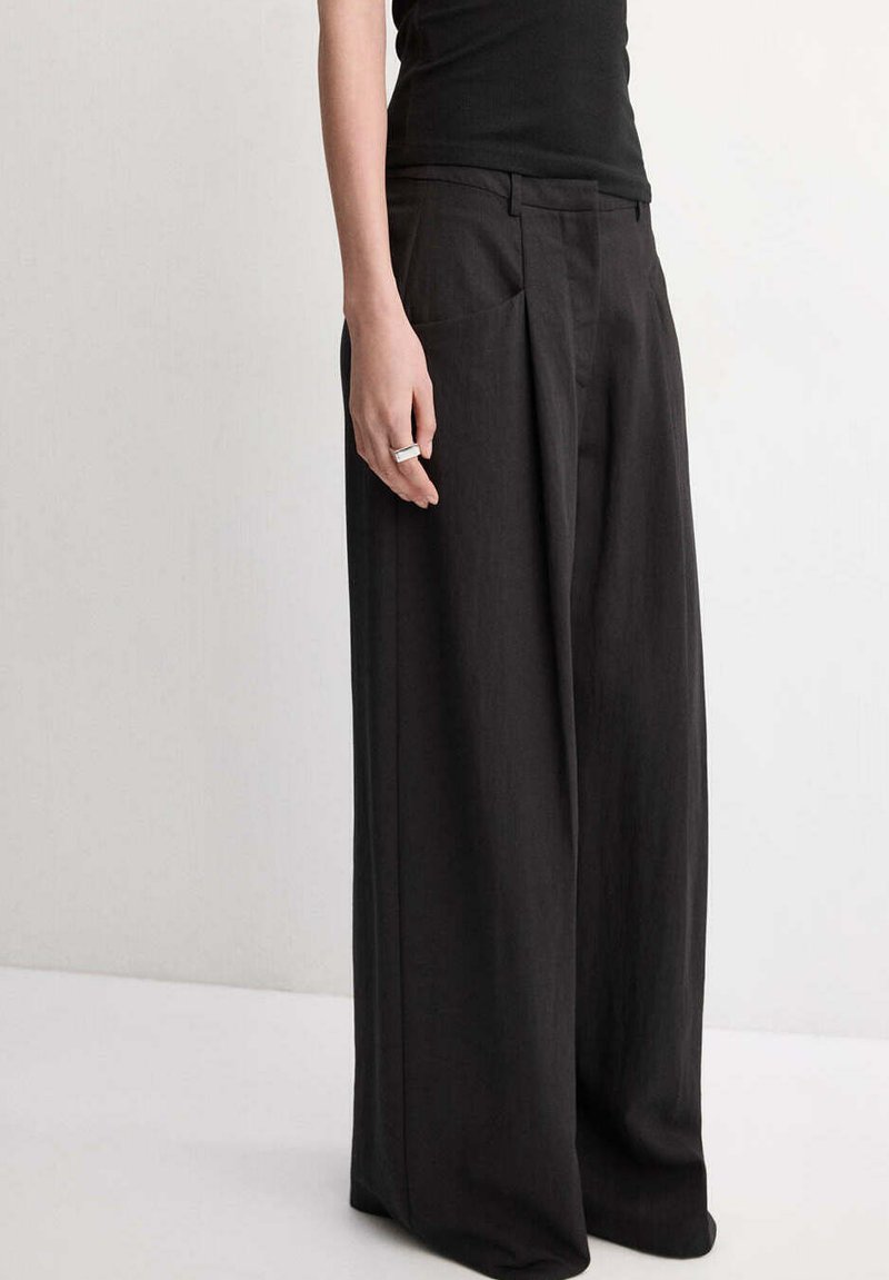 Personne portant un pantalon noir à jambes larges et un haut noir sans manches, avec une bague argentée à la main gauche.