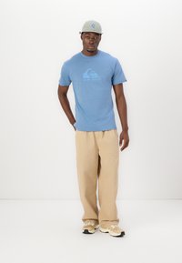 Blå bomulds T-shirt med et bølge-logo, parret med beige vidde-bukser og lyse sneakers. En lysegrå kasket fuldender outfittet.