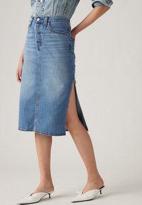 Falda midi de mezclilla azul claro con corte recto, abertura frontal y bolsillos traseros. Combinada con mules blancos con un detalle de lazo.