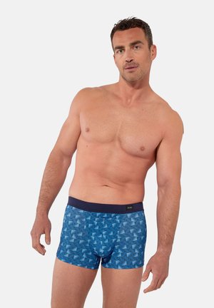 Oberkörperfreier Mann steht, trägt blaue gemusterte Boxershorts mit dunklem Bund, vor schlichtem hellgrauem Hintergrund.