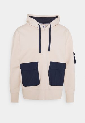Haut à capuche zippé beige avec des accents de poche bleu marine, cordons de serrage et texture douce. Dispose de deux grandes poches avant pour la fonctionnalité.