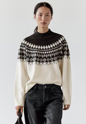 Kvinde iført en sort og hvid mønstret strikket sweater og sorte jeans, der holder en lille sort håndtaske mod en ensfarvet baggrund.