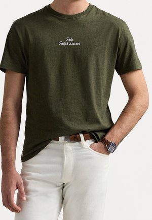 Homme portant un t-shirt Polo Ralph Lauren vert olive rentré dans un pantalon blanc avec une ceinture marron et une montre-bracelet à la main gauche.