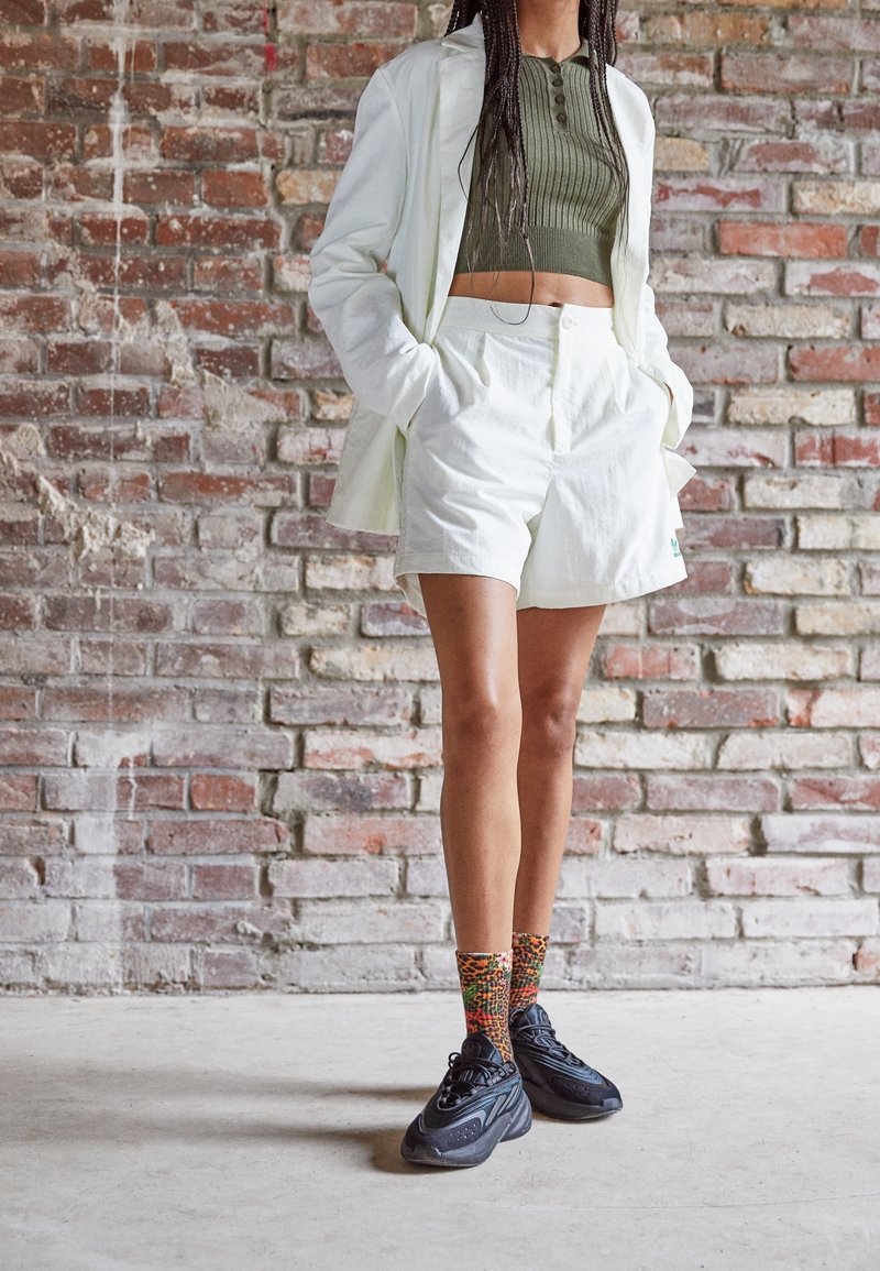 Valkoinen bleiseri oliivinvihreän ribattun crop-topin päällä, valkoiset räätälöidyt shortsit, mustat lenkkarit ja värikkäät kuviolliset sukat. Rakeinen tiiliseinä taustalla.