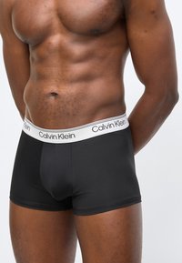 Ανδρικός κορμός που φοράει μαύρα εσώρουχα τύπου μπόξερ Calvin Klein με λευκή μέση και λογότυπο, τα χέρια χαλαρά πίσω από την πλάτη, σε απλό ανοιχτό φόντο.