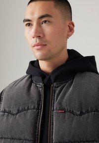 Sivi trapezasti prsluk od jeansa s zlatnim patentnim zatvaračem, crna dukserica ispod, i mali crveni Levi's logo na džepu na prsima. Glatka tekstura.