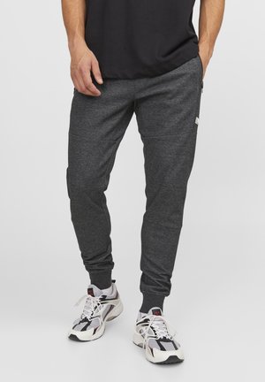 Homme portant un pantalon de jogging gris foncé et des chaussures de sport blanches, noires et rouges, debout avec une main dans la poche devant un fond blanc.
