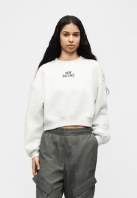 Γυναίκα με μακριά σκούρα μαλλιά, φοράει ελαφρύ γκρι κοντομάνικο φούτερ New Balance και σκούρο γκρι παντελόνι cargo, στέκεται με το ένα χέρι στην τσέπη.