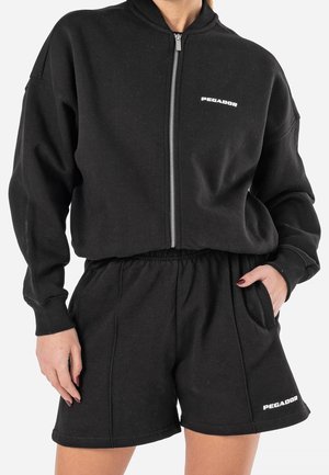Vrouw draagt een zwart sweatvest met ritssluiting en bijpassende korte broek, beide met het witte logo "PEGADOR", staand met één hand in de zak.