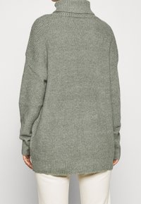 Suéter de punto oversize en verde apagado, con un alto cuello de tortuga, textura acanalada, hombros caídos y un dobladillo trasero ligeramente más largo.