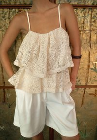 Top in pizzo bianco con volant sovrapposti e spalline sottili, abbinato a pantaloni corti plissettati bianchi. Lo sfondo presenta un motivo floreale.